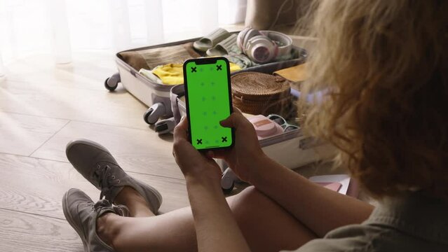 Smartphone with green chroma key mockup in hand of woman. Green screen phone, suitcase ready to go. Woman scrolls down feed curated travel recommendations in mobile app, advertisement. Weekend getaway