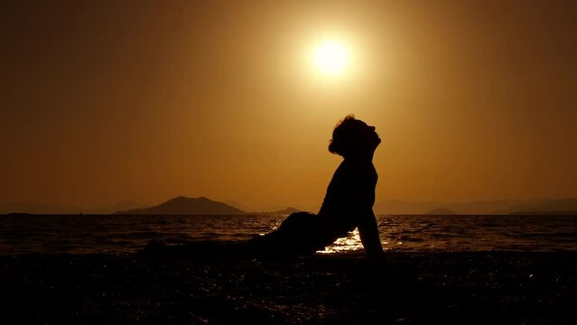 Woman doing stretching asana on dusk. A woman silhouette lie down on the shore and stretching her back and neck against bright sun during nightfall time.