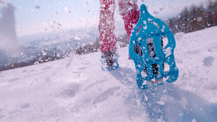 LOW ANGLE VIEW, CLOSE UP: Lady in blue snowshoes and snow flying behind her