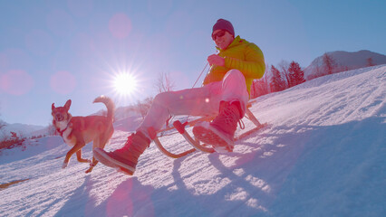 CLOSE UP, LENS FLARE: Cheerful man sledding and brown dog running by his side
