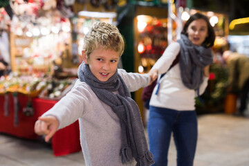 Obraz premium Portrait of tween boy pointing to desired thing and pulling mothers hand during walk at festive fair on Christmas Eve