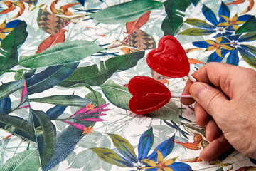 man's hand holding two red heart-shaped lollipops with a tropical pattern background.