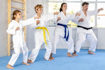 Fototapeta na wymiar In gym, family of four repeats movements of karate card and practicing kicks and punches in unison, is intently preparing for sparring.
