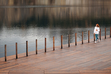 Concentrated woman with red hair in white hoodie and blue leggings walking on wooden pier near the lake after jogging. Runner holds bottle of water and phone in her hands
