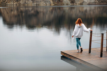 Fototapeta na wymiar Sporty woman with red hair in white hoodie and blue leggings standing at wooden pier near the lake and enjoying peaceful view. Fittness woman holding phone and bottle. Back view