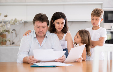 Saddened father reads studies financial expenditure documents. Thoughtful man is encouraged and hugged by wife and children. Woman with husband carefully reads sad news in financial business report