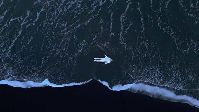Dramatic view of desperate male wearing white clothes lying motionless on black sanded beach while wild foaming waves strike the shore, washing the man. High quality 4k footage