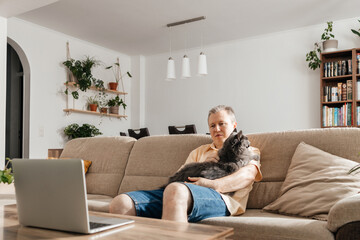 A man with a gray cat in his arms sits on the sofa and looks at his laptop. Remote learning or work, relaxation after work with your favorite pet