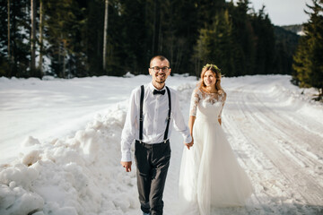 The bride and groom are running along a snowy road against the background of a pine forest and beautiful contrast sunlight. Side view. Winter wedding. Place for logo.