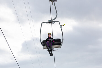 Smiling young woman in ski outfit sitting alone on chairlift, raising to slopes for skiing on background of cloudy winter sky, bottom view
