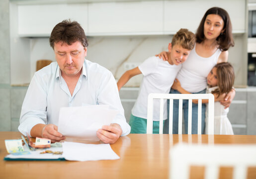 Wistful Father Counting Money Looking Through Papers Sitting In The Kitchen, His Wife And Children Looking At Him With Curiosity Standing Behind