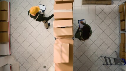 Top down view of warehouse logistics coordinators examining cardboard box parcels in parallel, comparing findings. Distribution center coworkers making sure stocks and inventory are well organized