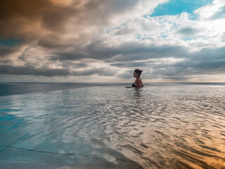 Portrait of a woman standing in an infinity pool looking over the sea at sunset