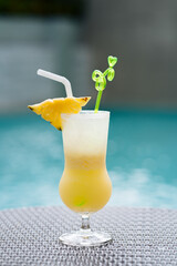 A tropical cocktail in a tall glass with a yellow-to-white gradient, garnished with star-shaped pineapple and a green swizzle stick, against a blurred pool background, indicating a relaxed daytime.