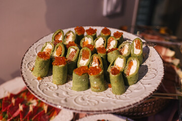 Festive appetizers on receptions. Spinach crapes rolls with caviar, salmon and cream cheese inside. Plate with savoury seafood pancake rolls with roe. Banquet table with healthy food at restaurant.