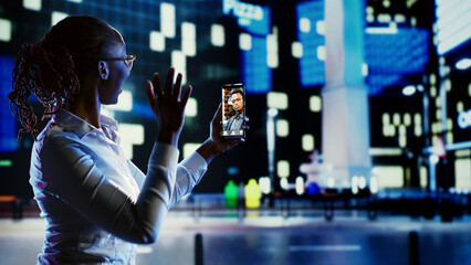 Woman greeting husband in online videocall while strolling around city streets at night. Person...