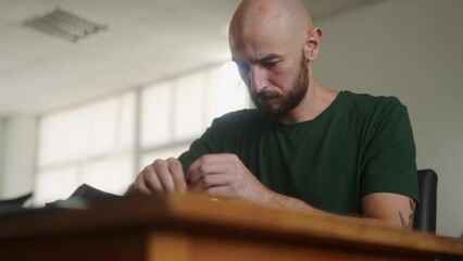 Professional leather craftsman at work table making stitching holes in black handbag with leather punch and hammer. Bold man with beard in khaki T-shirt sewing purse in workshop
