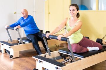 Fototapeta premium Smiling young woman performing set of pilates exercises on reformer in fitness studio..