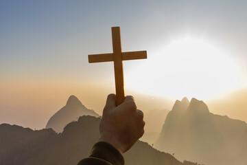 human hands praying to the GOD while holding a crucifix symbol with bright sunbeam on top mountains