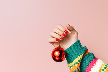Christmas bauble hangs on woman's hand with red manicure against pink background.
