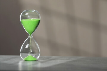 Hourglass with green flowing sand on table against light grey background, space for text