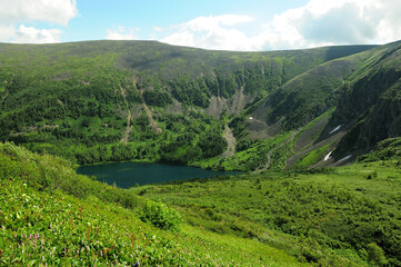 Fototapeta na wymiar A beautiful small lake in a deep intermountain basin with snow on the slopes and banks under a sunny summer sky.