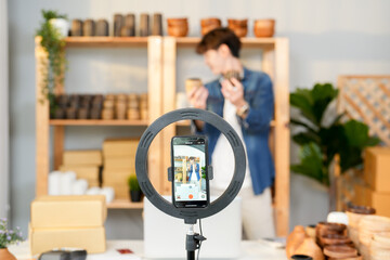 Asian business owner man live streaming handmade product and review compare feature design while selfie record clip video with mobile and ring light for promote online e-commerce in studio apartment - obrazy, fototapety, plakaty