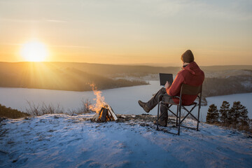  man freelancer sitting on snow mountain top using laptop working remotely at sunset. man tourist resting near campfire using laptop.