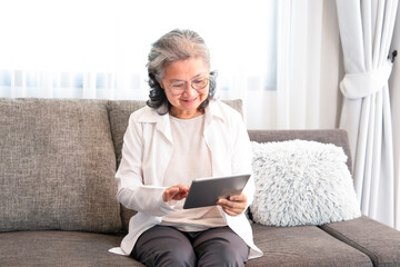 asian senior woman sitting on couch,relaxing in the living room,using internet online on tablet...