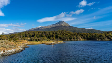 Beautiful blue lake. Yellowed grass and thickets of trees grow on the shore. A picturesque mountain against a blue sky and clouds. Argentina. Lapataia Bay. Tierra del Fuego National Park. Ushuaia