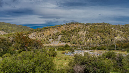 Fototapeta na wymiar Landscape of Patagonia. The river winds through the valley. The twisted trunks of withered trees are visible on the shore. Wooded mountains against a blue sky and clouds. Argentina. Tierra del Fuego 