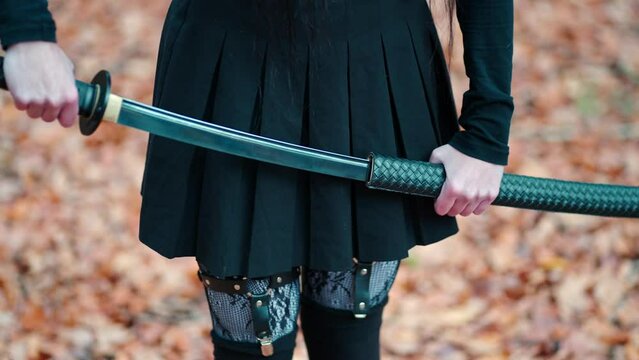 Young girl in a black skirt with garters and stockings pulls out a katana sword. Preparing for battle. On the background of colorful autumn leaves. Shot of a shiny sword blade