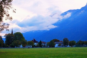 Fototapeta na wymiar Landscape of Interlaken in the morning; composed of lawn, houses and buildings amid trees, on mountain with cloudy-sky background, Switzerland