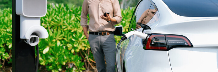 Young man use smartphone to pay for electricity at public EV car charging station green city park....