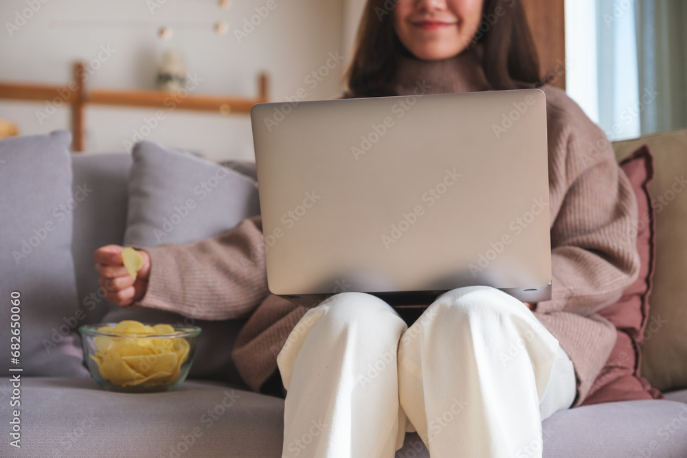 Wall mural Closeup image of a young woman eating potato chips while using laptop at home