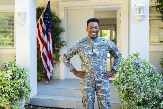 Happy african american male soldier with hands on hips outside home with usa flag, copy space