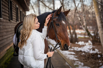 Fototapeta premium Young beautiful rider woman blonde with long hair in white clothes standing near horse