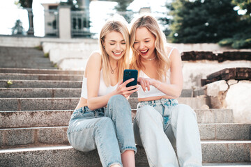 Two young beautiful smiling trendy female. Carefree women posing in street. Positive models having fun. Cheerful and happy. Hold smartphone, look at mobile cellphone screen, use phone apps