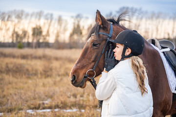 Beautiful blond professional female jockey standing near horse in field. Friendship with horse