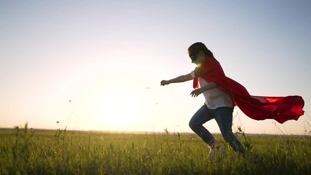 Happy child girl in superhero costume in the park. A girl in a mask runs through the grass in a field at sunset. Concept of success and victory. Dream girl is actively playing in the park outdoors.