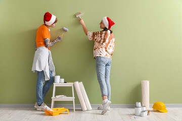 Young pregnant couple with Santa hats and painting tools near green wall, back view