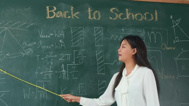 Back to school concept. Asian female teacher smiling with wooden stick pointing to blackboard at school in classroom, Happy beautiful young woman standing hold pointer to back board