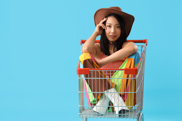 Beautiful young stylish Asian woman sitting in shopping cart on blue background