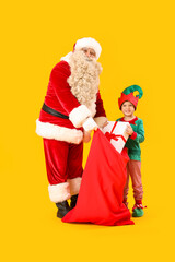 Santa Claus and cute little elf with bag full of Christmas gifts on yellow background