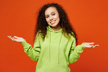 Young sad doubtful woman of African American ethnicity she wear green hoody casual clothes shrugging shoulders looking puzzled spread hands isolated on plain red orange background. Lifestyle concept. - obrazy, fototapety, plakaty