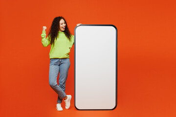 Full body young woman of African American ethnicity wear green hoody casual clothes big huge blank screen mobile cell phone smartphone with area do winner gesture isolated on plain orange background.