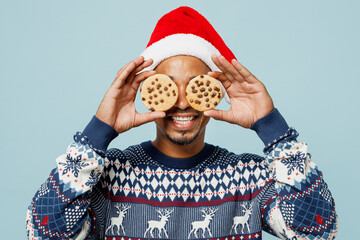 Young fun man wear knitted sweater Santa hat posing cover eyes with two chocolate biscuits cookies...