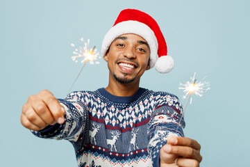 Young smiling man wears knitted sweater Santa hat posing hold in hands two bengali fires look camera isolated on plain pastel blue background Happy New Year 2024 celebration Christmas holiday concept