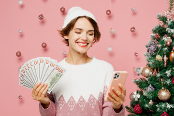 Merry young woman wears white sweater hat posing hold mobile cell phone fan of cash money in dollar...