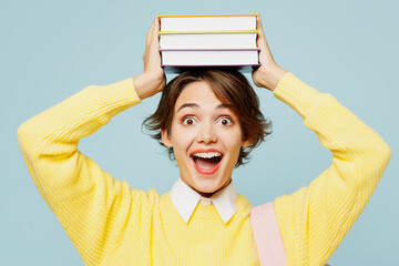 Close up young surprised clever fun woman student wear casual clothes yellow sweater backpack bag hold stack of books on head isolated on plain blue background. High school university college concept. - obrazy, fototapety, plakaty